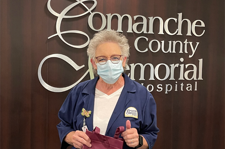 Mary Wilson, an older white woman with short grey hair and glasses, posing in front of the CCMH logo wall